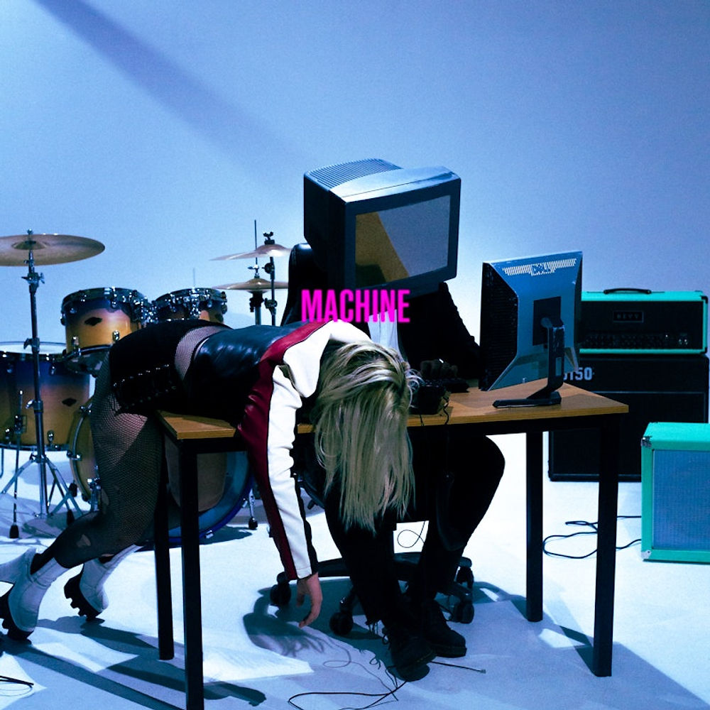 A person leaning over a desk, appearing exhausted, next to someone with a vintage TV head. Text "MACHINE" is visible. Drum set in background.
