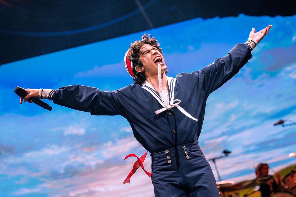 Performer in a navy sailor suit with arms outstretched, holding a mic. Background shows a cloudy sky. Energetic and joyful mood.
