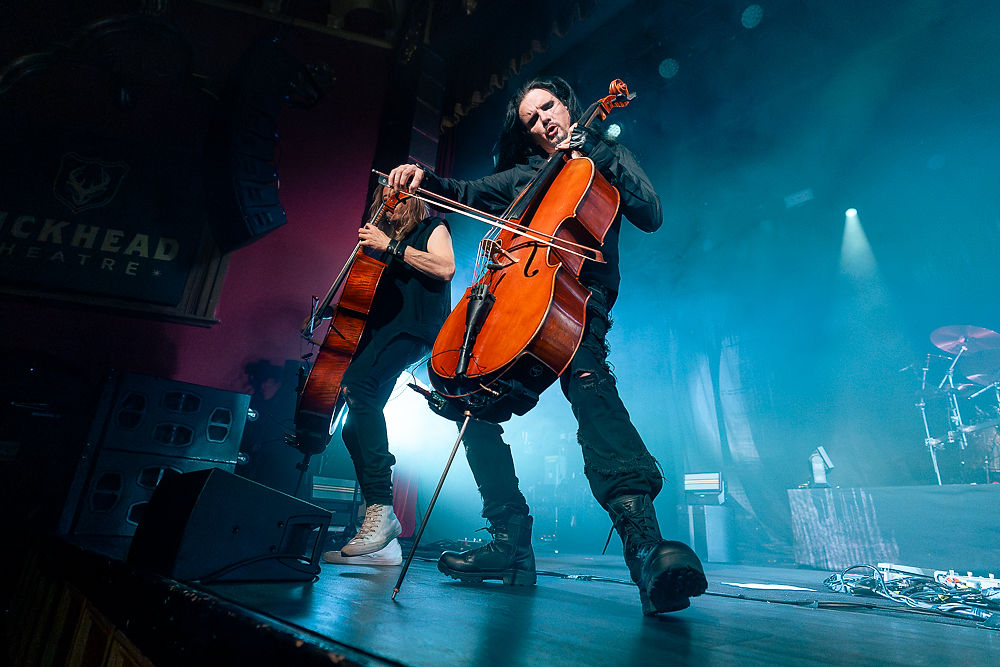 Cellists perform energetically on stage, intense expression, blue-lit background, "BUCKHEAD THEATRE" visible, lively concert atmosphere.