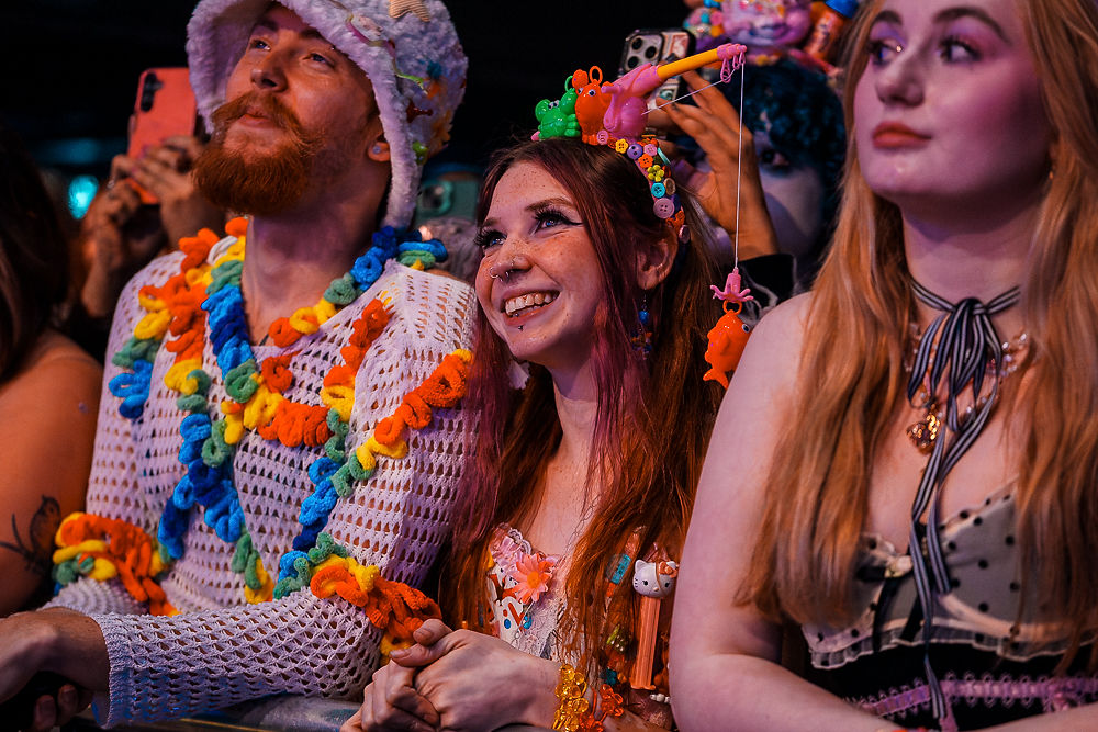 People in colorful outfits and headpieces smile and watch a performance. The setting is energetic, with vibrant colors and an excited mood.