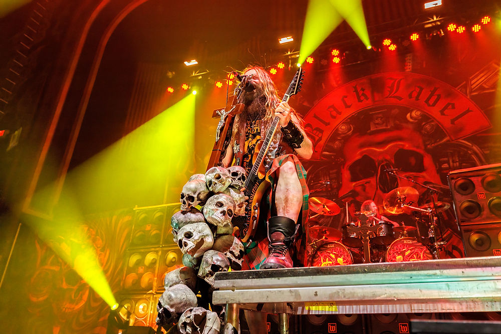 Rock musician plays guitar on stage, surrounded by skull props and yellow lights. "Black Label" visible in red backdrop. Intense energy.
