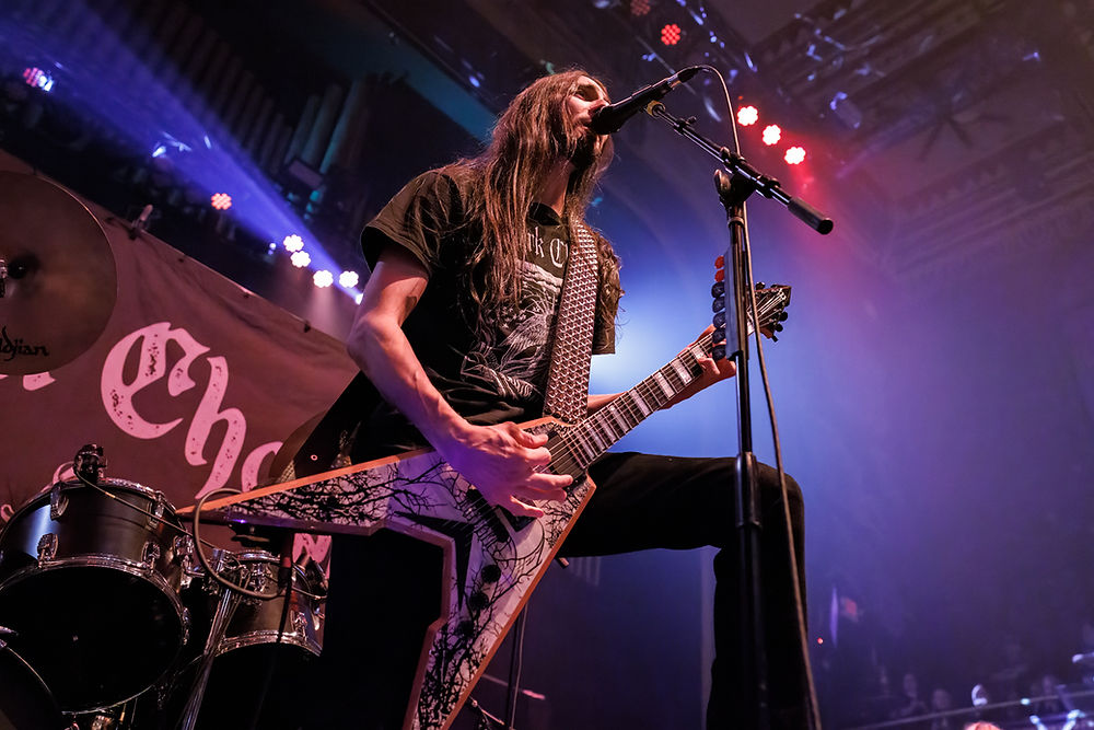 Guitarist with long hair performs passionately on stage, holding a guitar with an intricate design. Colorful stage lights illuminate the scene.