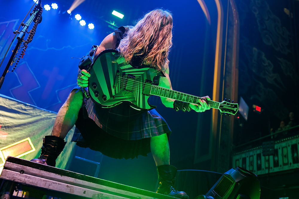 Guitarist with long hair, wearing plaid skirt, plays guitar on stage. Green and blue lights highlight energetic performance, rock setting.