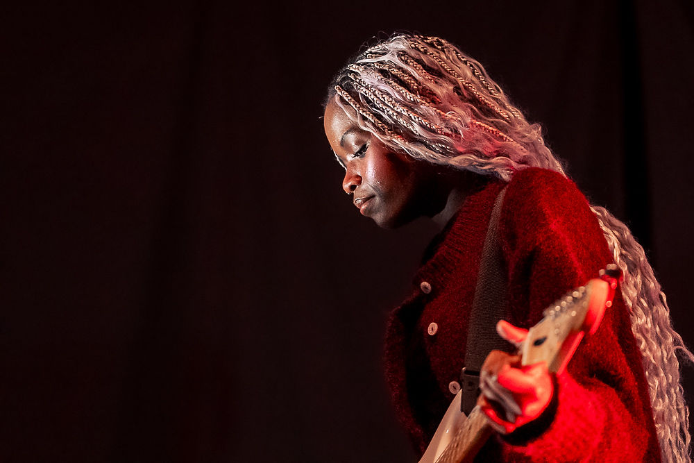 Musician with long braided hair plays electric guitar against a dark background, wearing a red sweater, focused and immersed in music.