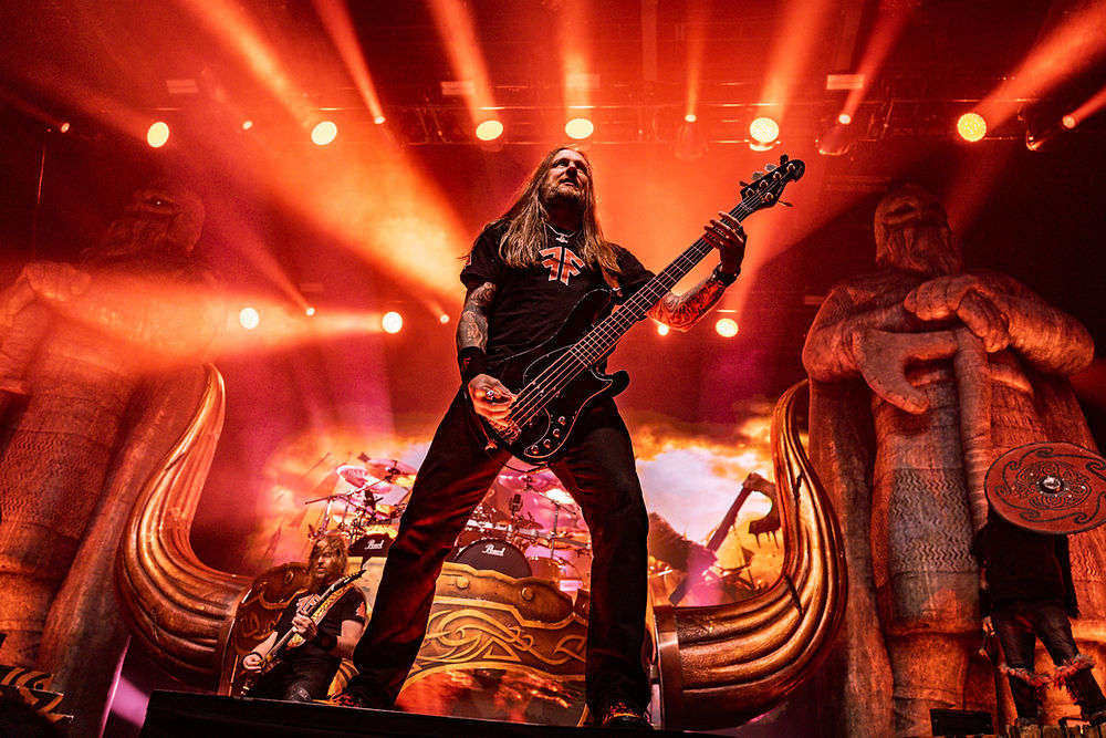 Bassist performs on stage with red lighting, flanked by large Viking statues. Dramatic atmosphere with instruments and intense energy.