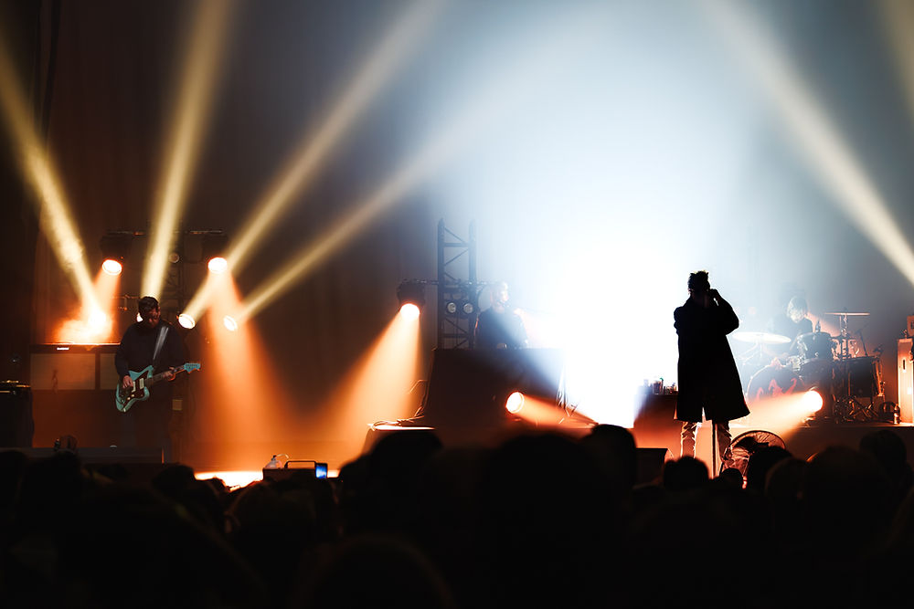 Silhouetted band performs on a bright concert stage with orange and white spotlights, guitarist left, singer center, drummer right.