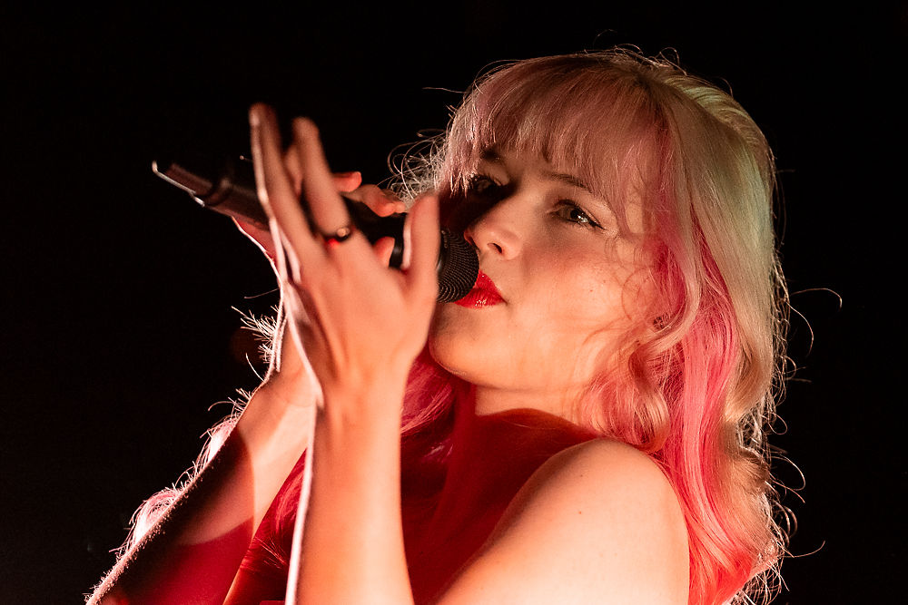 Singer with pink hair and red lipstick passionately performing on stage, holding a microphone against a dark background.