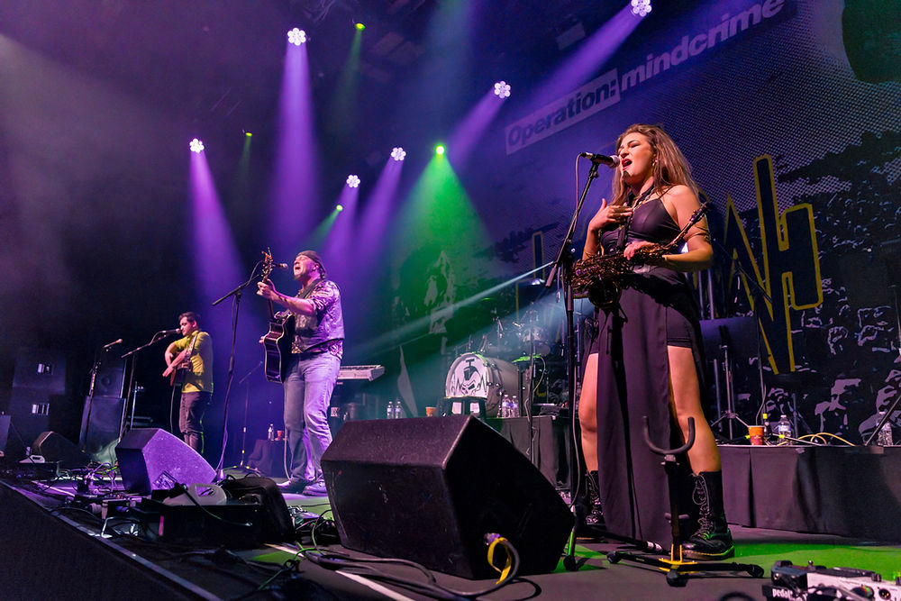 Musicians perform energetically on stage with instruments under purple and green lights. "Operation: mindcrime" is visible in the background.