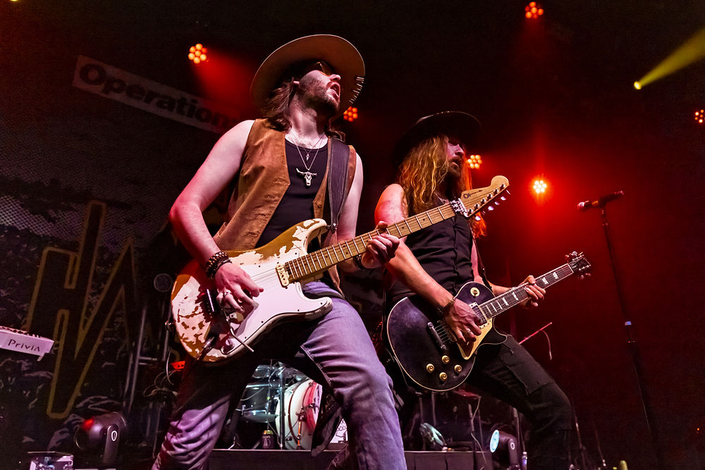 Two guitarists in cowboy hats perform energetically on stage under red lights, with a drum set and banners in the background.