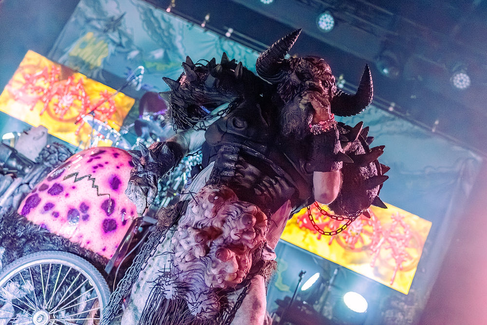 A performer in a spiked, horned costume sings on stage. Bright screens and colorful lighting create an energetic concert vibe.