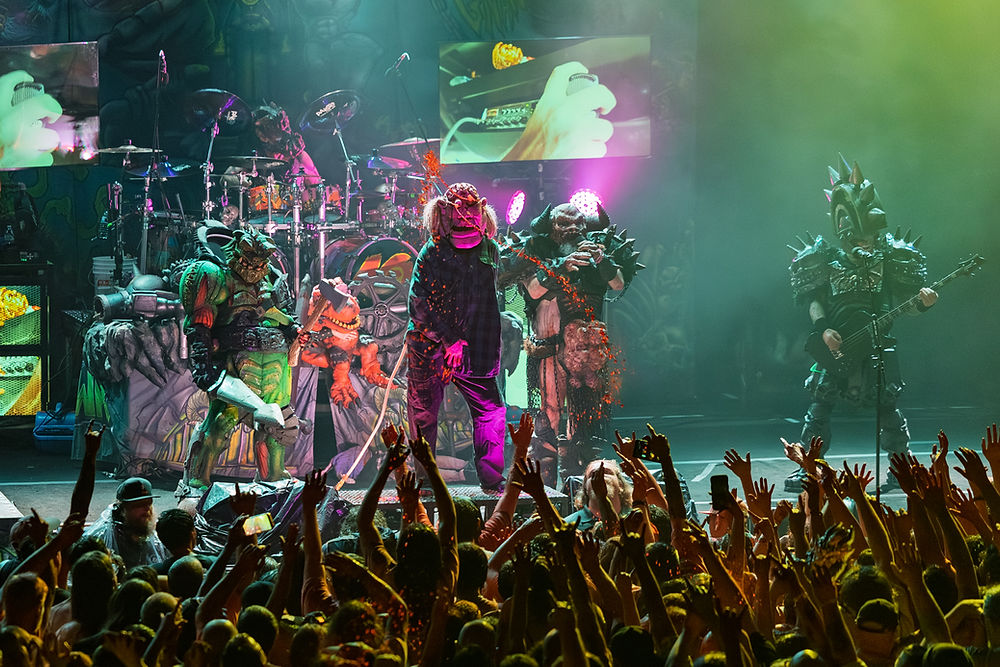 Band in elaborate costumes perform on stage amid colorful lights and screens. Crowd raises hands, creating an energetic concert atmosphere.