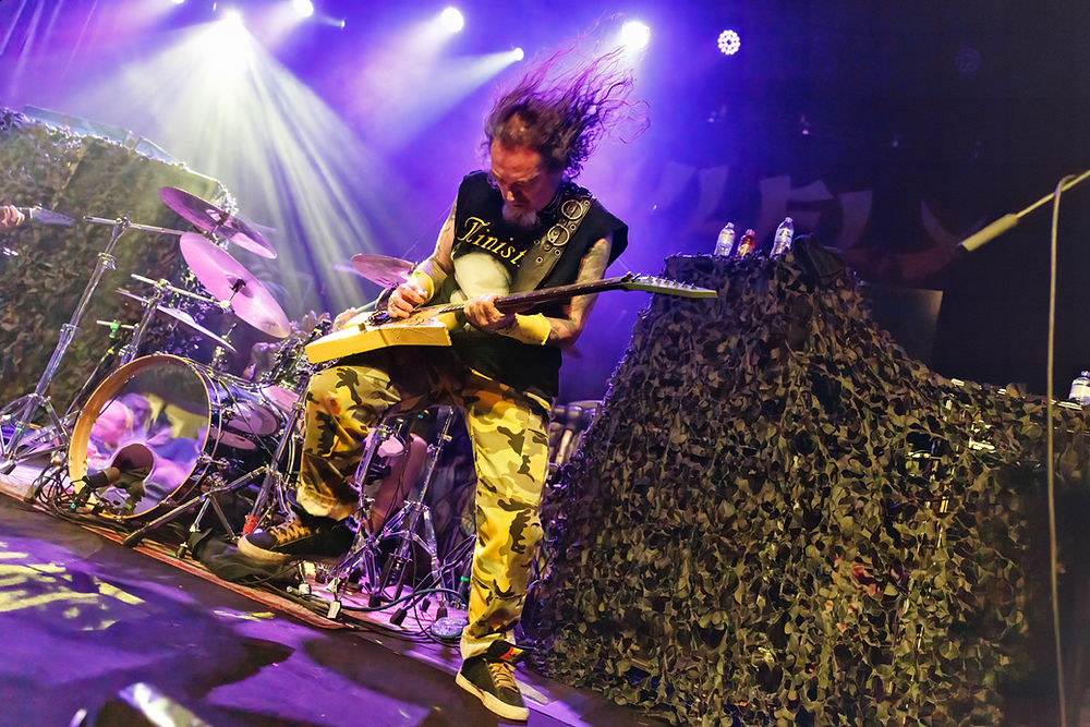Guitarist in camo pants plays energetically on stage with purple lights. Hair flying, drums nearby, camo-covered equipment. Rock concert vibe.