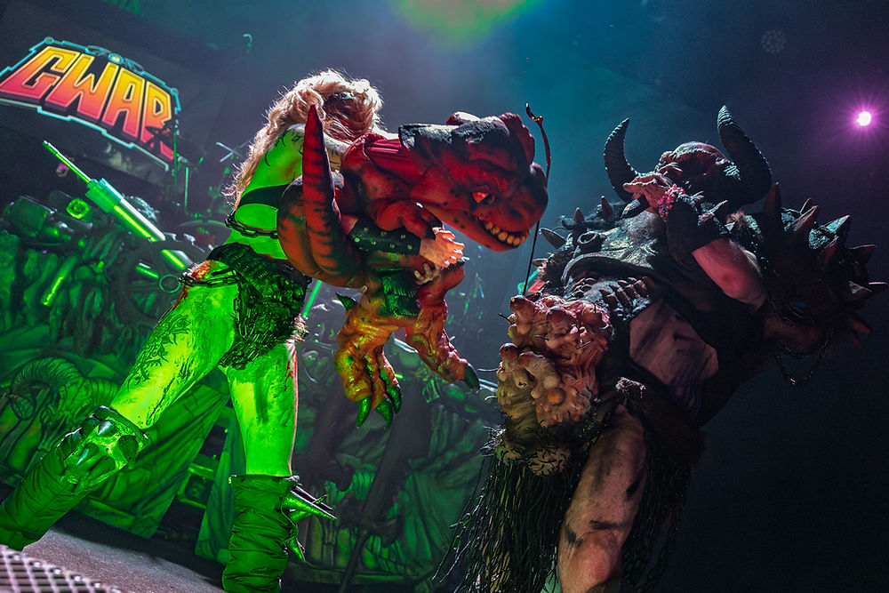 Performers in elaborate monster costumes face off on stage, under vibrant green and purple lights. "GWAR" sign in the background.