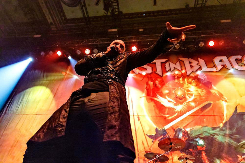 Singer in black leather performs passionately on stage, pointing forward. Colorful backdrop with "IN BLACK" text and dramatic lighting.