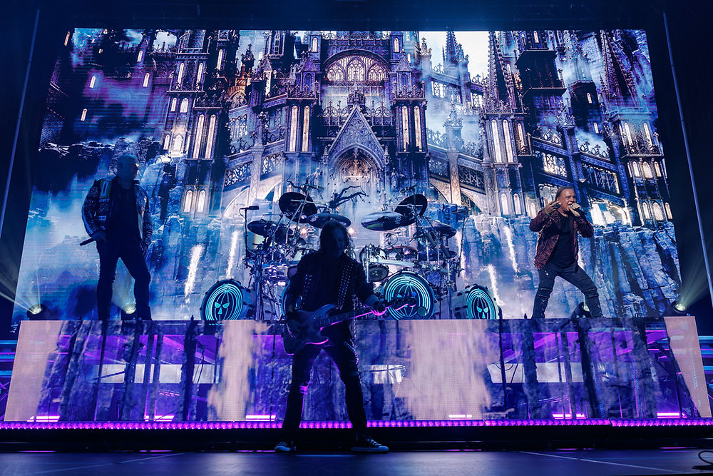 Rock band performing on stage with a Gothic castle backdrop. Vibrant purple lighting, energetic atmosphere, musicians playing instruments.