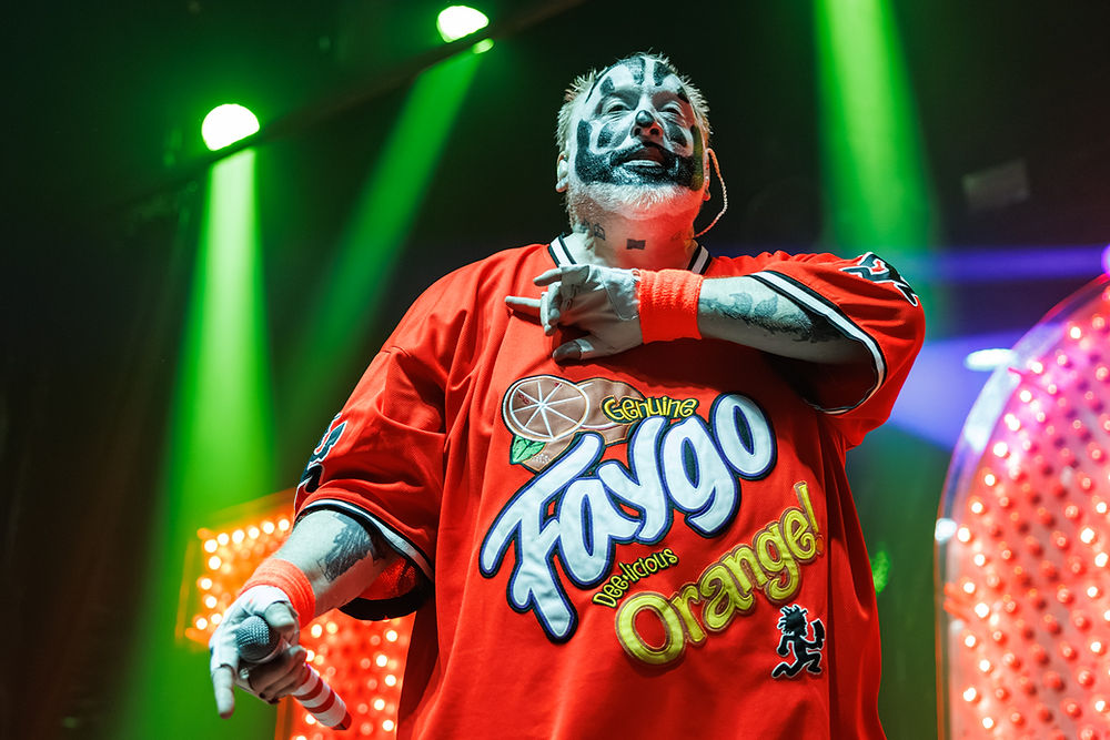 Masked performer in red Faygo Orange shirt sings onstage under green lights, holding a microphone.