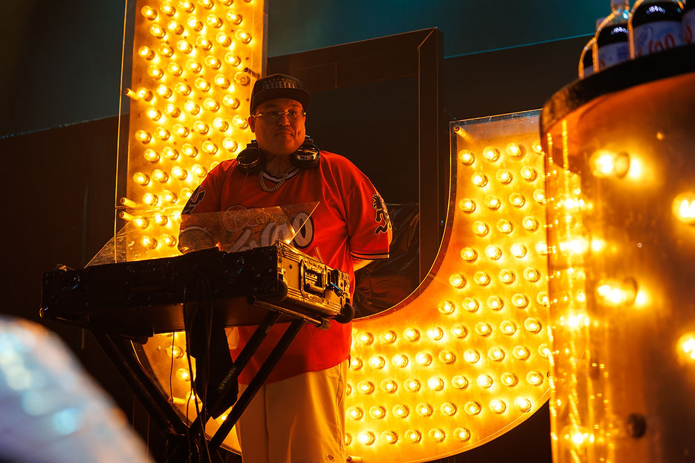 DJ in red shirt and cap stands behind turntables, framed by glowing yellow marquee lights in a dark club.