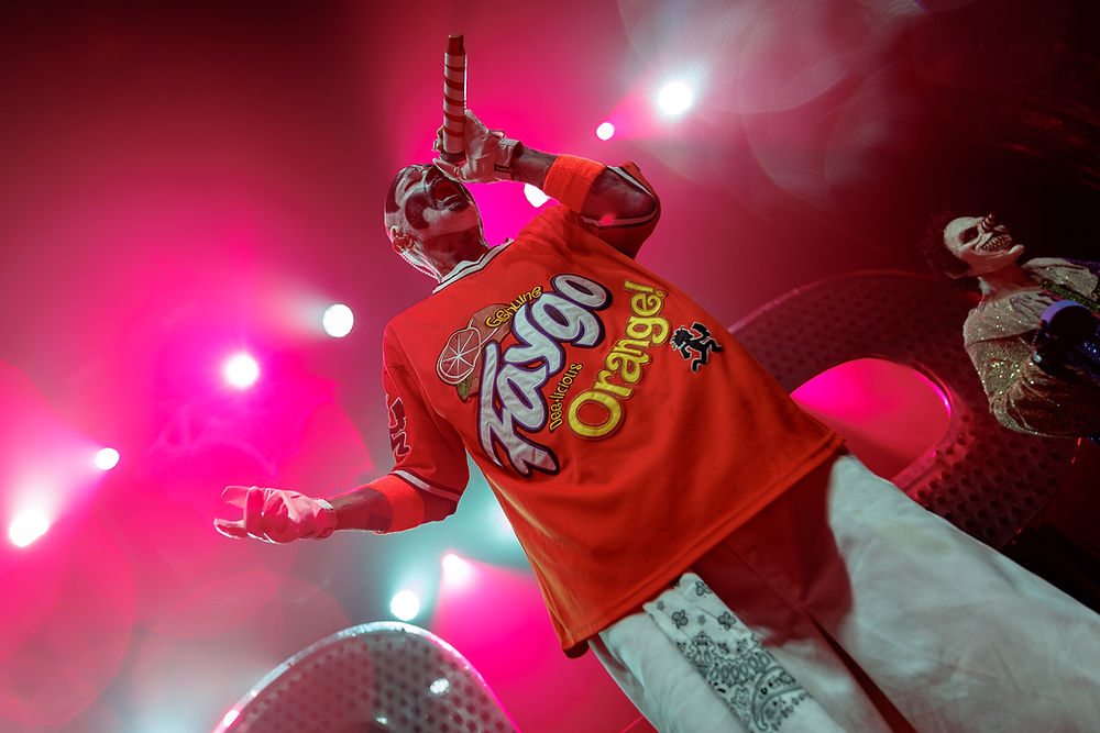 Orange-clad stage performer sings into a striped mic under pink lights; Fanta Orange logo on shirt, masked dancer at right.