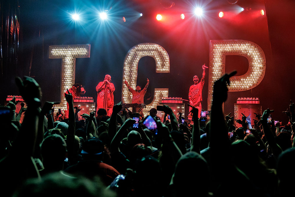 Clowns on a concert stage spray the crowd in front of giant lit letters T C P, with hands raised and phones recording.