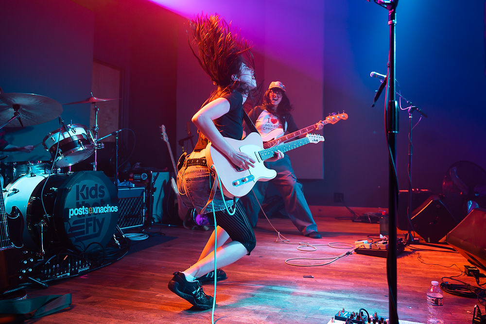 Two performers on stage; one plays electric guitar energetically, hair flipping, and the other plays bass. Drum set labeled Kids. Vibrant lights.