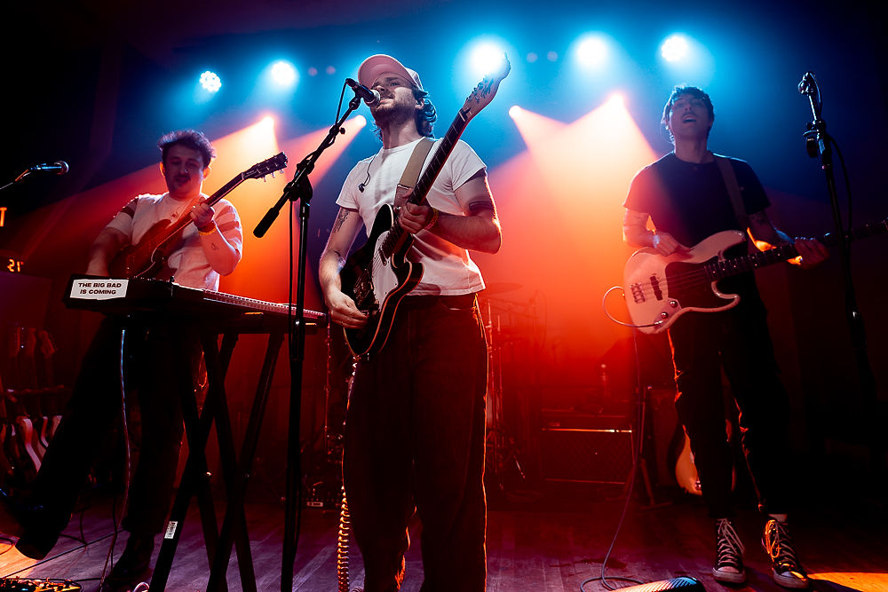 Band performing on stage with vibrant red-blue lighting. Three musicians play guitar and keyboards. Text on keyboard reads "The Big Bad is Coming."