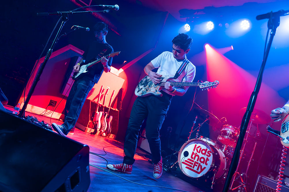 Band performing on stage with colorful lights. Two guitarists play energetically. Red drum kit features "Kids That Fly" logo. Mood is lively.