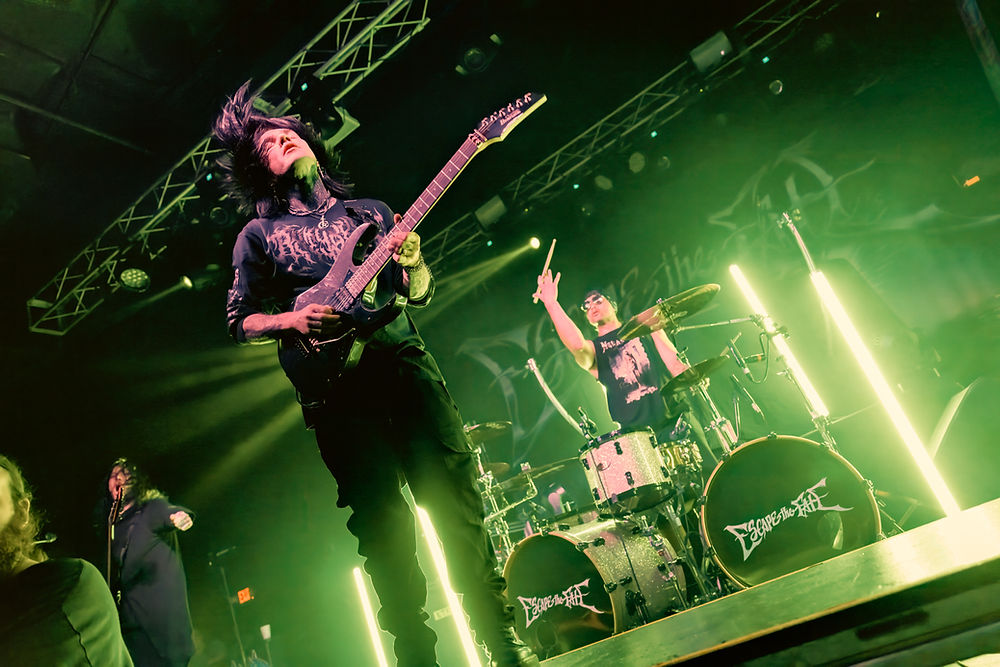 Band performing on stage with intense green lighting. Guitarist and drummer in action, "Escape the Fate" visible on drums, energetic mood.