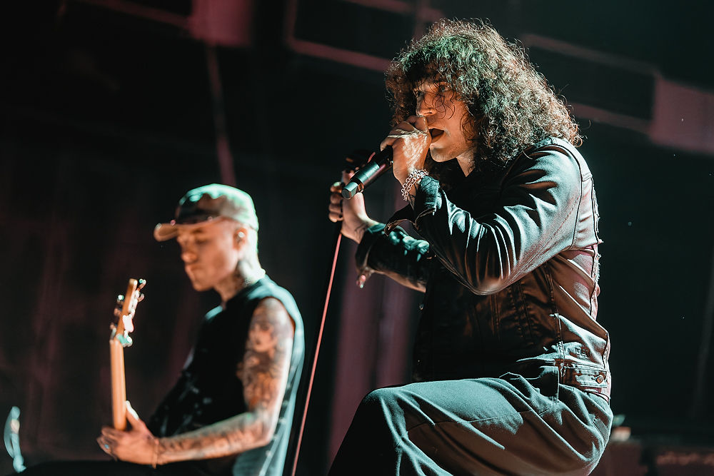 Singer with curly hair sings into a microphone onstage while a tattooed guitarist plays beside him under warm stage lights.