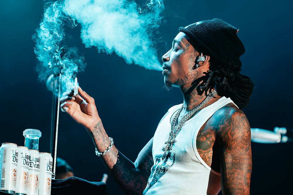 Tattooed man in black beanie exhales smoke on a dark blue stage; Liquid Death cans sit nearby.