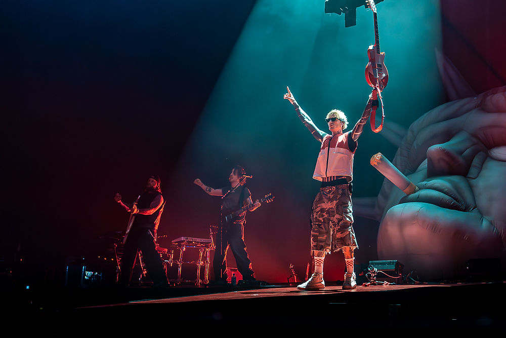 Rock band performs on a dark stage; singer raises both arms under teal and red lights, with a giant hand and cigarette backdrop.