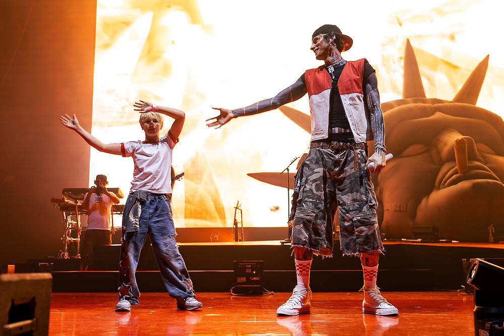 Two performers on a lit concert stage, arms outstretched, with a bright orange screen and band gear behind them.