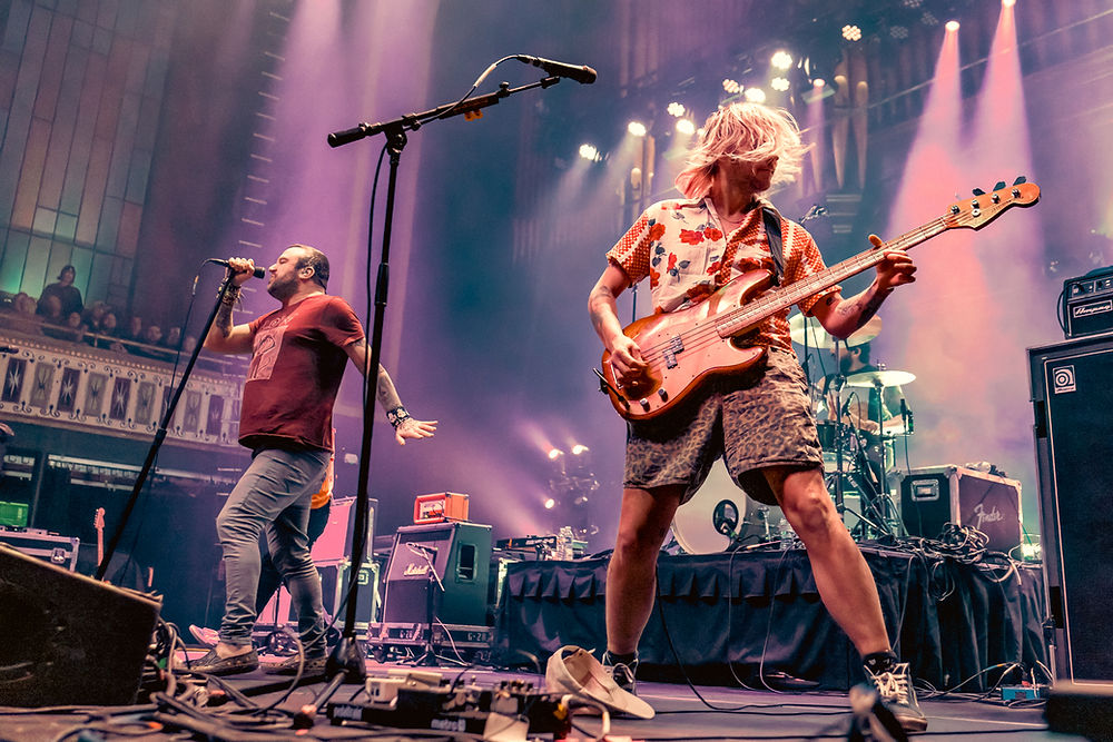 Musicians performing energetically on stage with colorful lighting. One sings into a mic, the other plays bass. Audience and equipment in the background.