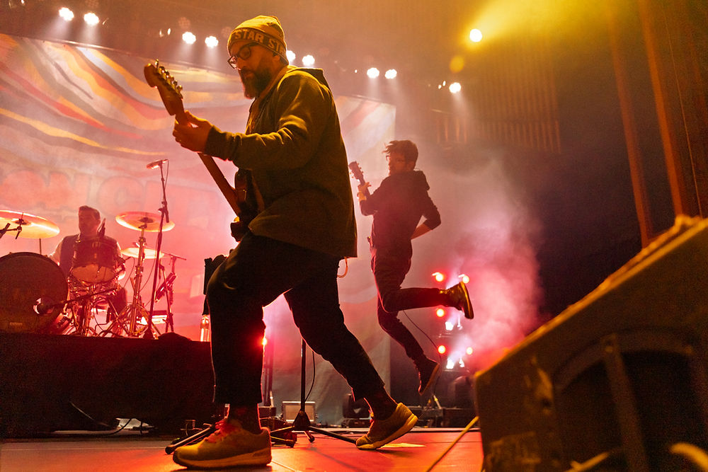 Musicians perform energetically on stage with colorful lights and smoke. One jumps mid-air, another plays guitar. Mood is lively and dynamic.