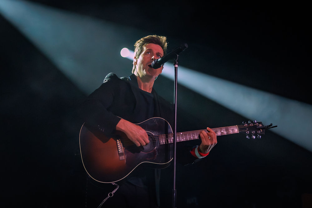 A person plays guitar and sings into a microphone on stage, lit by a spotlight. The dark background enhances the dramatic mood.