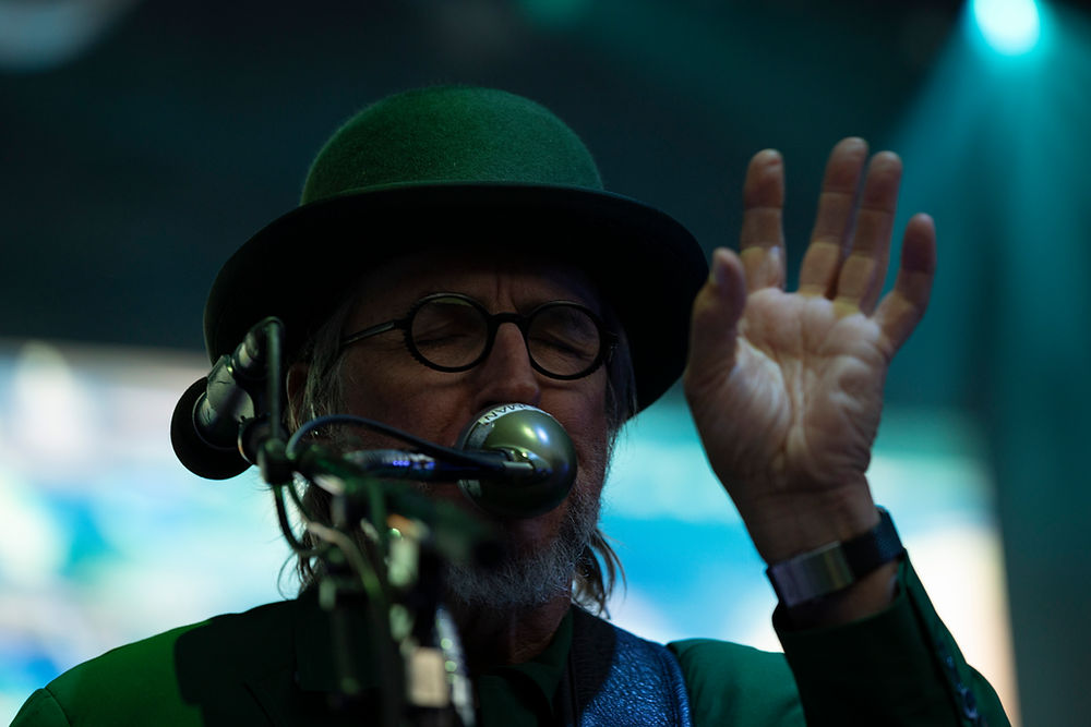 Older man in a green hat and glasses sings into a microphone with one hand raised under blue-green stage lights.