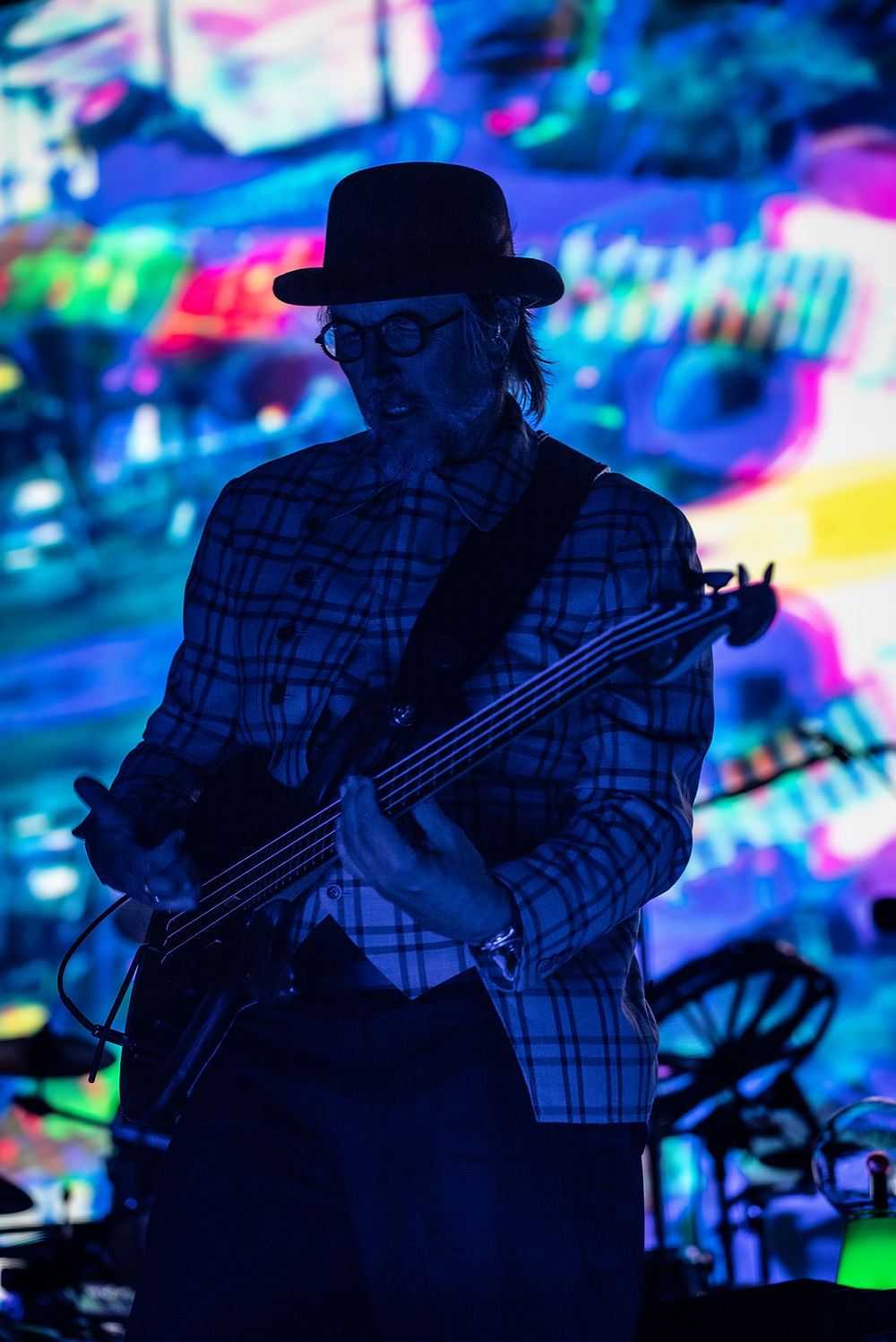 Silhouetted bassist in a hat and plaid shirt plays onstage before a vivid blue, pink, and green abstract light backdrop.