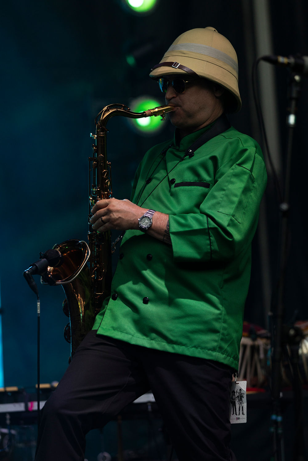 Saxophonist in a green jacket and tan helmet-style hat plays on a dim stage with glowing green lights, focused mood.