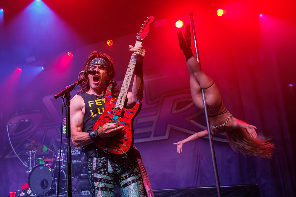 Rock guitarist with a red electric guitar and "Feel Lucky" shirt performs energetically. Pole dancer in leopard print upside down. Red stage lights.