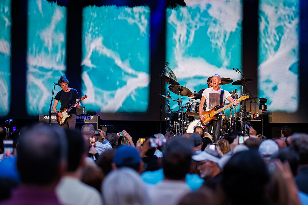 Musicians perform energetically on stage with guitars and drums. The background shows an ocean wave design. A crowd watches, capturing photos.