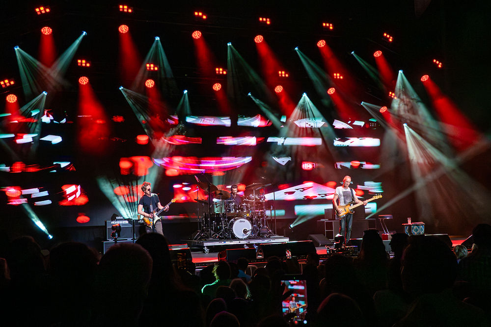 Three musicians perform on stage with vibrant red and blue lights and abstract visuals. The energetic atmosphere captivates the audience.