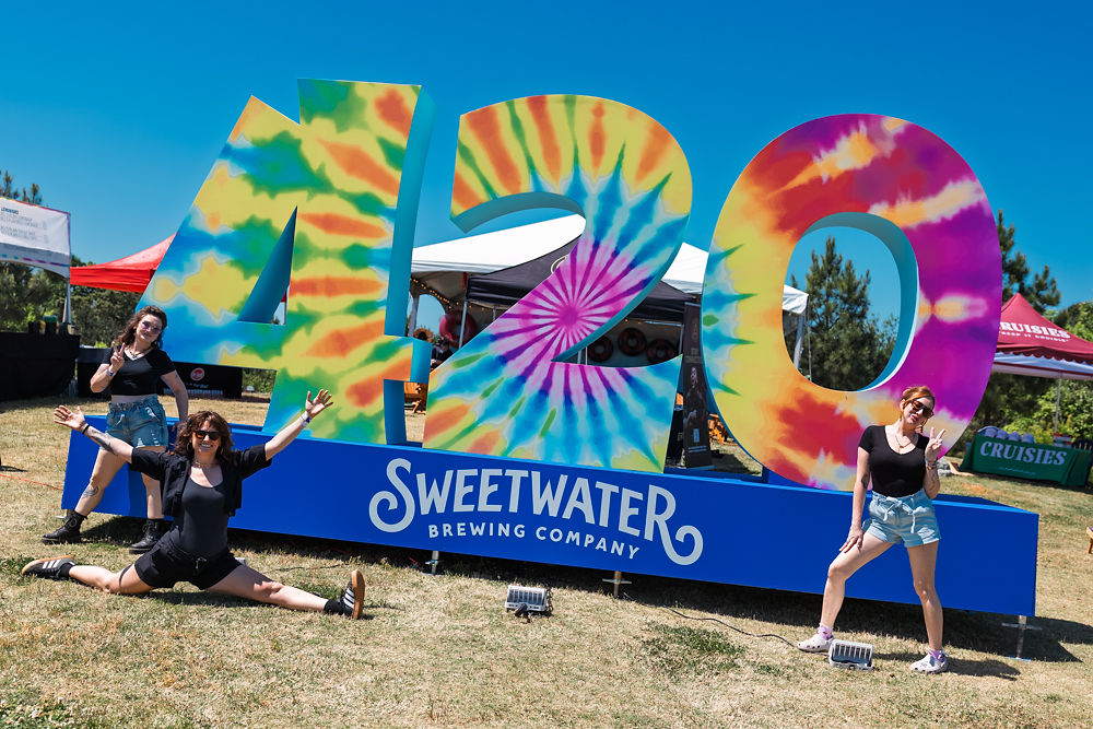 Three people pose joyfully in front of large, colorful "420" sign with "Sweetwater Brewing Company" text. Festival tents and trees in background.