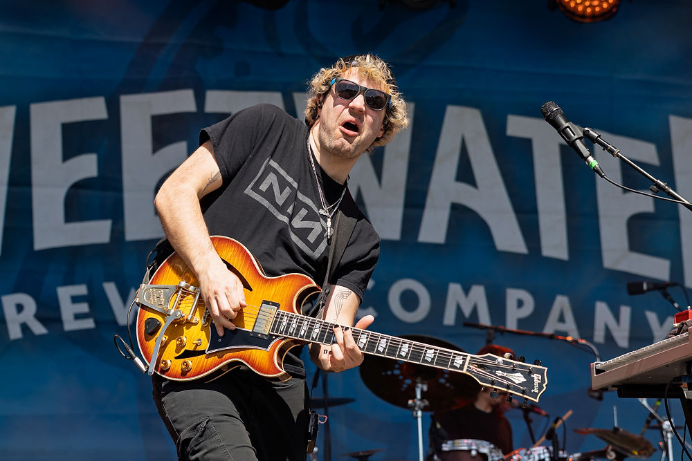 Musician playing guitar on stage with "Sweetwater" backdrop, wearing sunglasses and a black T-shirt. Daytime outdoor performance.