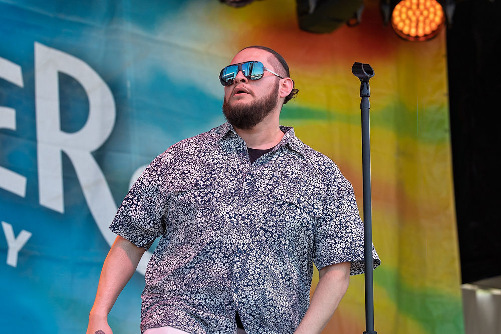 Singer in a patterned shirt and sunglasses stands by a mic on a colorful stage. Large text in the background partially visible.