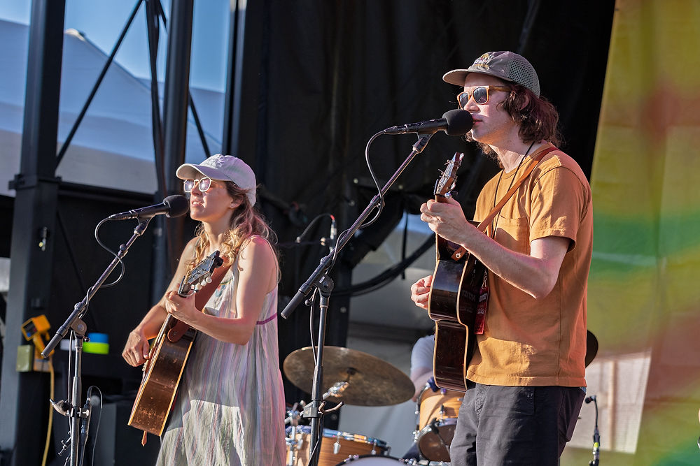 Two musicians with guitars sing on stage. Both wear caps and casual clothes. Drums in the background. Bright, lively atmosphere.