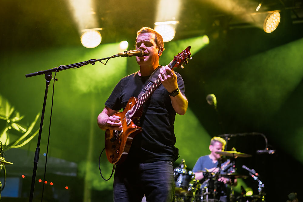 Musician sings and plays guitar on stage under bright lights. Drummer in background. Vivid green lighting and dynamic concert atmosphere.