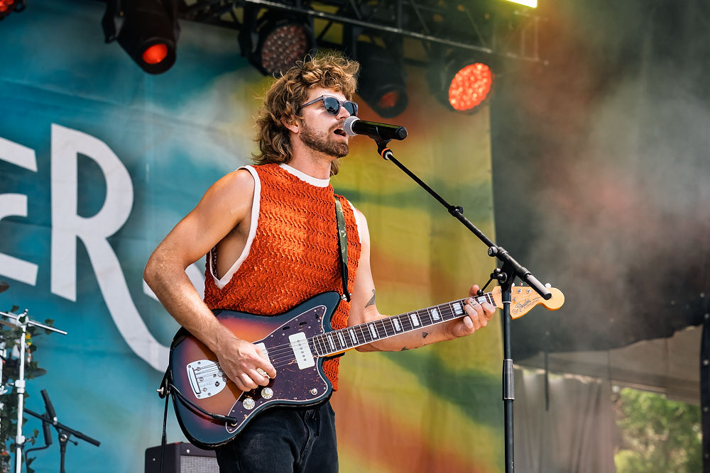 Musician in a sleeveless orange shirt plays guitar and sings on stage. Background shows colorful banner with partial text "ER".