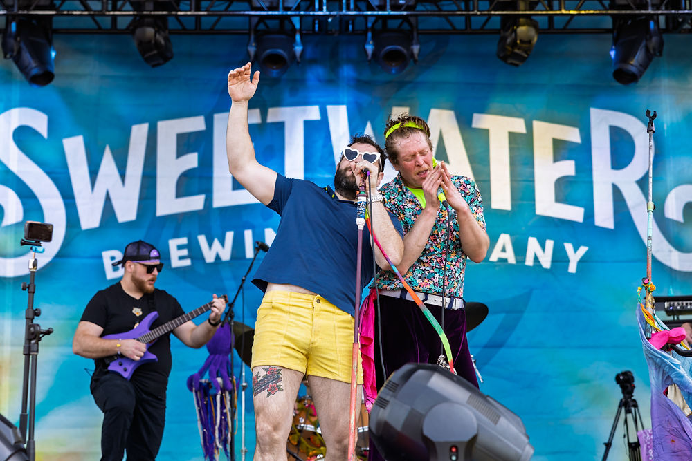 Two performers sing on stage, one in yellow shorts and heart glasses, the other in vibrant clothes. "Sweetwater Brewing Company" backdrop. Energetic mood.