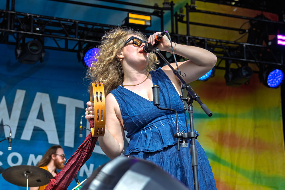 A woman with curly hair sings into a microphone, holding a tambourine on stage. She wears a blue dress, with colorful lights in the background.