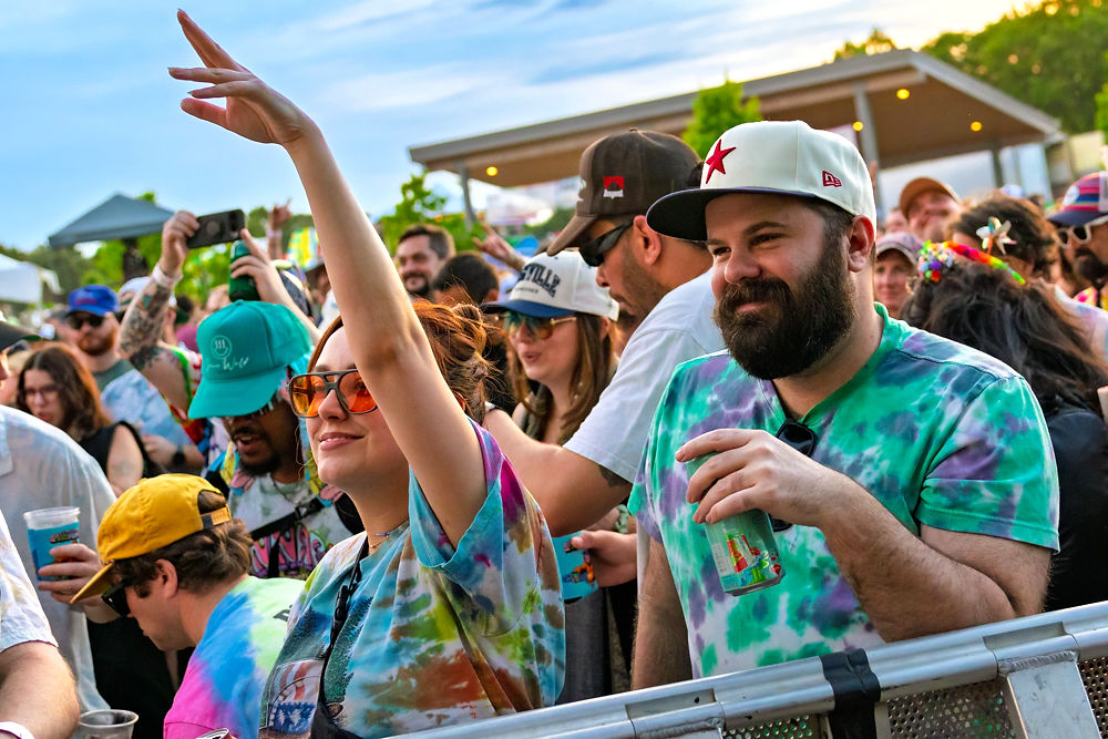People in colorful tie-dye outfits and sunglasses dance joyfully outdoors at a festival. A woman wears a flower in her hair. Bright, sunny day.