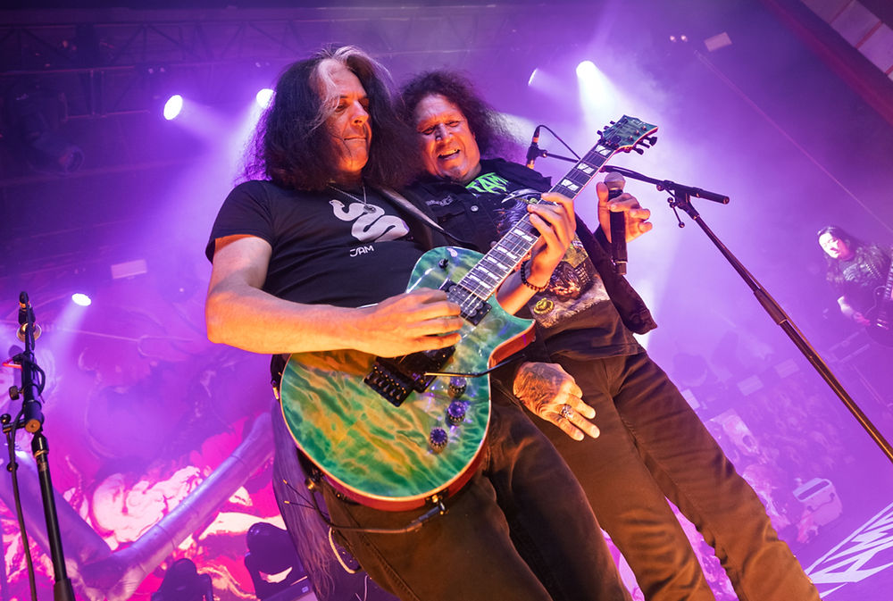 Musicians perform energetically on stage with bright purple lighting. One plays a turquoise guitar, audience and equipment in the background.